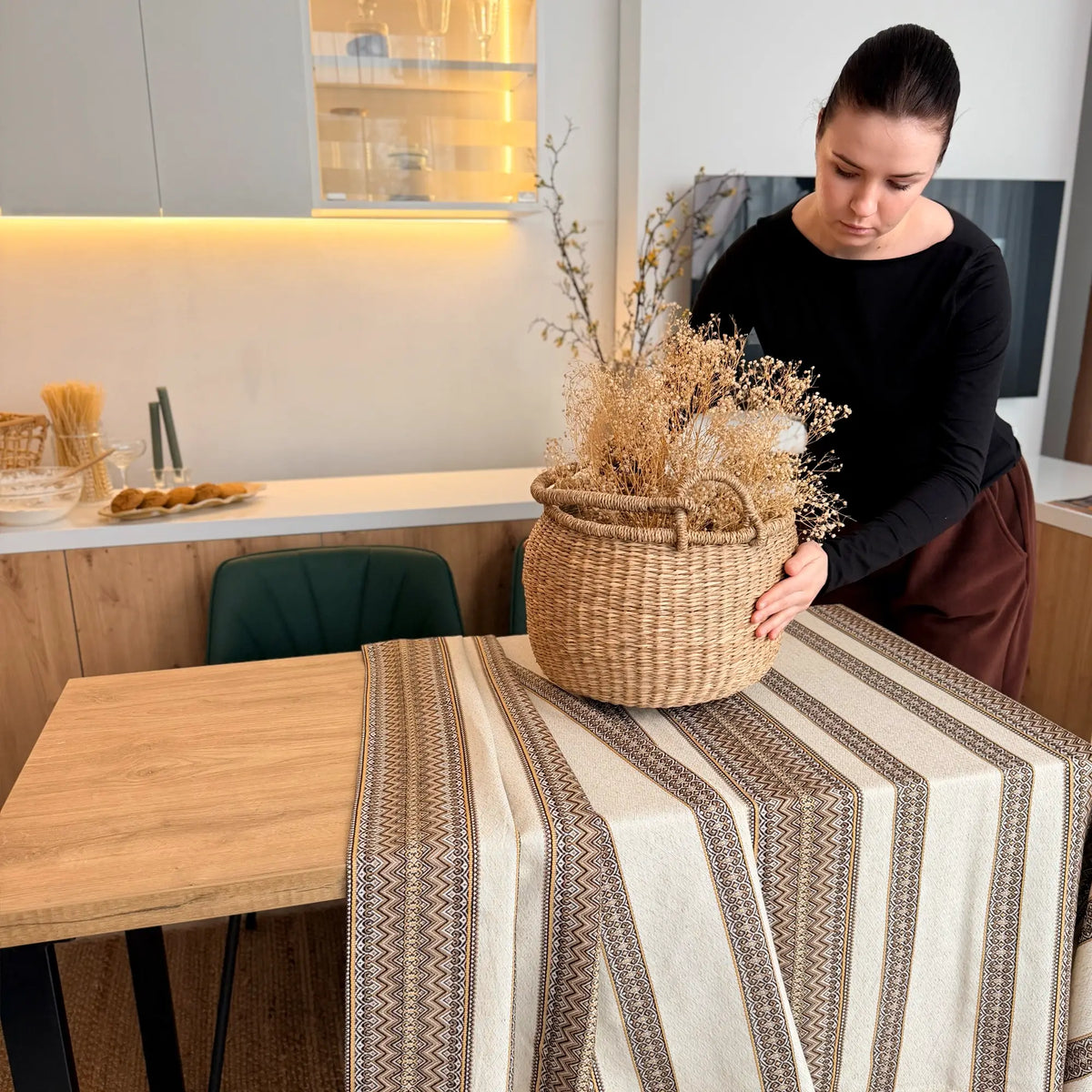 Woman arranging a woven basket with dried plants on a table in a kitchen.