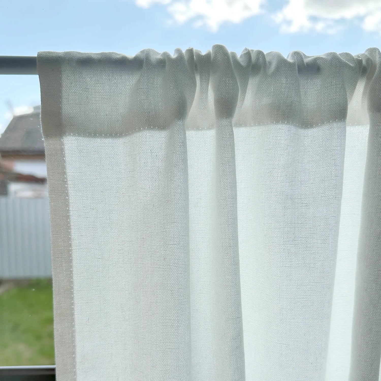 Sheer white curtain with a textured pattern on a rod against a blue sky.