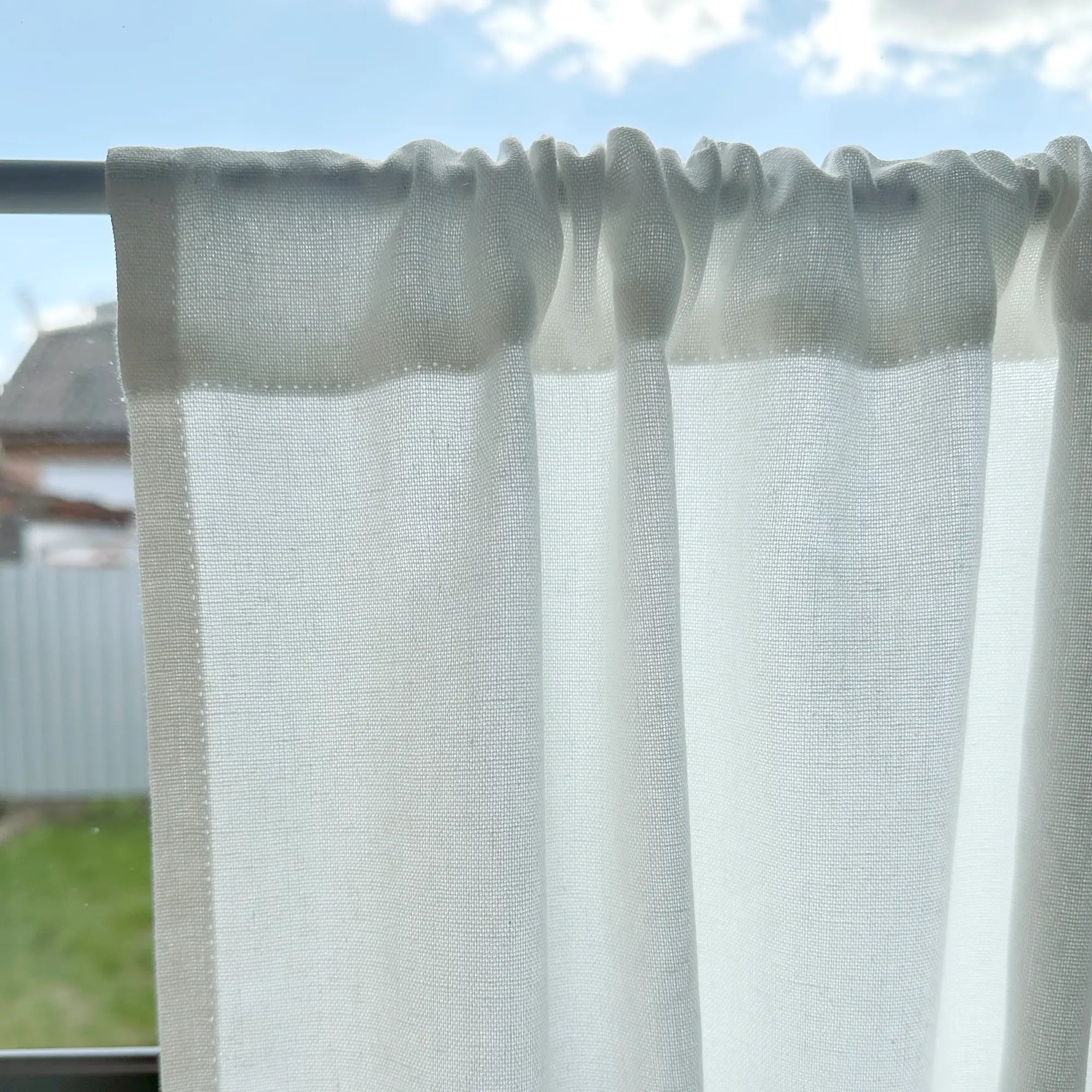 Sheer white curtain with a textured pattern on a rod against a blue sky.
