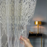 Decorative sheer curtain with intricate patterns held by a hand, blurred interior background