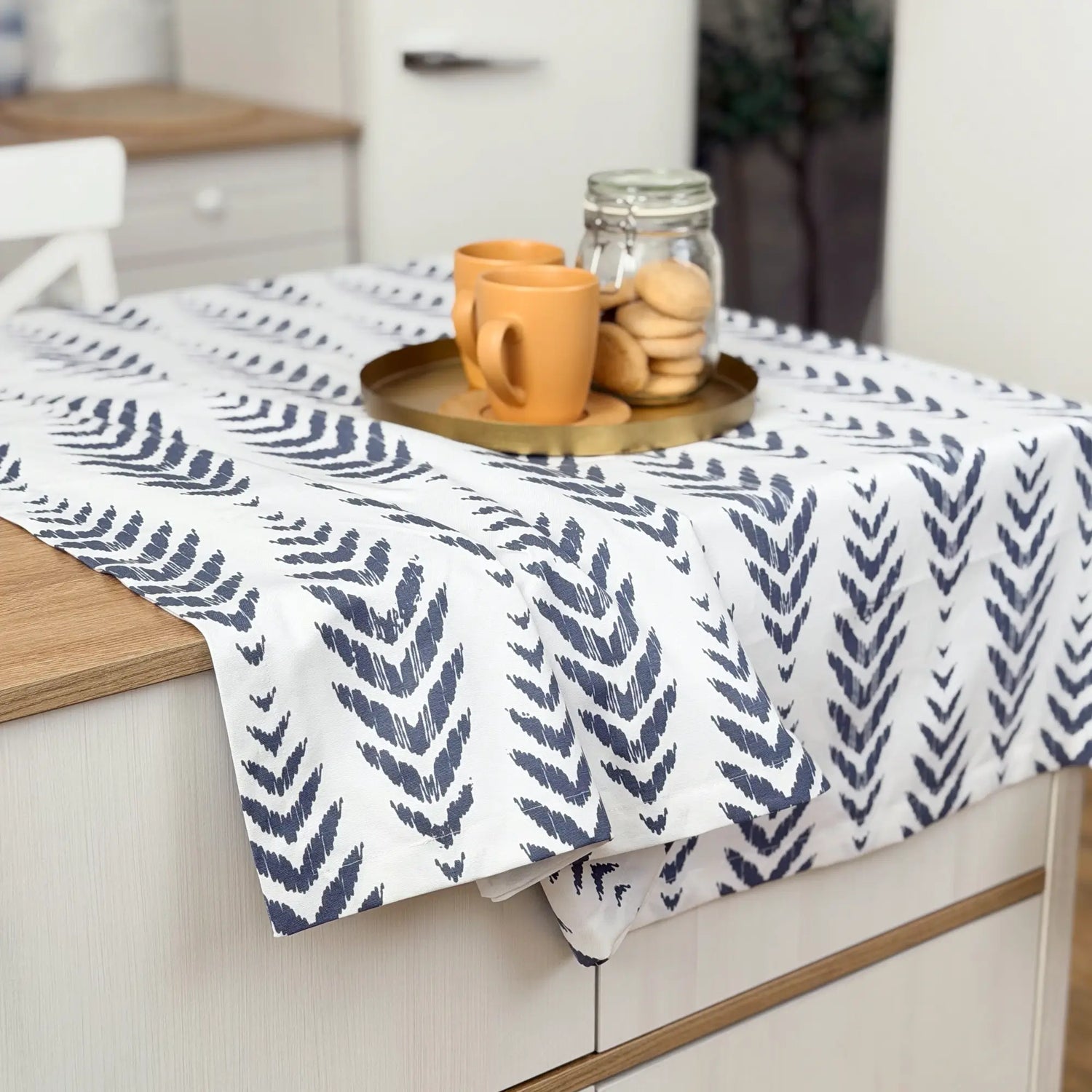 Table with a blue leaf patterned tablecloth, orange mugs, and cookies on a wooden surface.