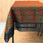 Tablecloth with red and green geometric pattern and black fringe on a wooden table.