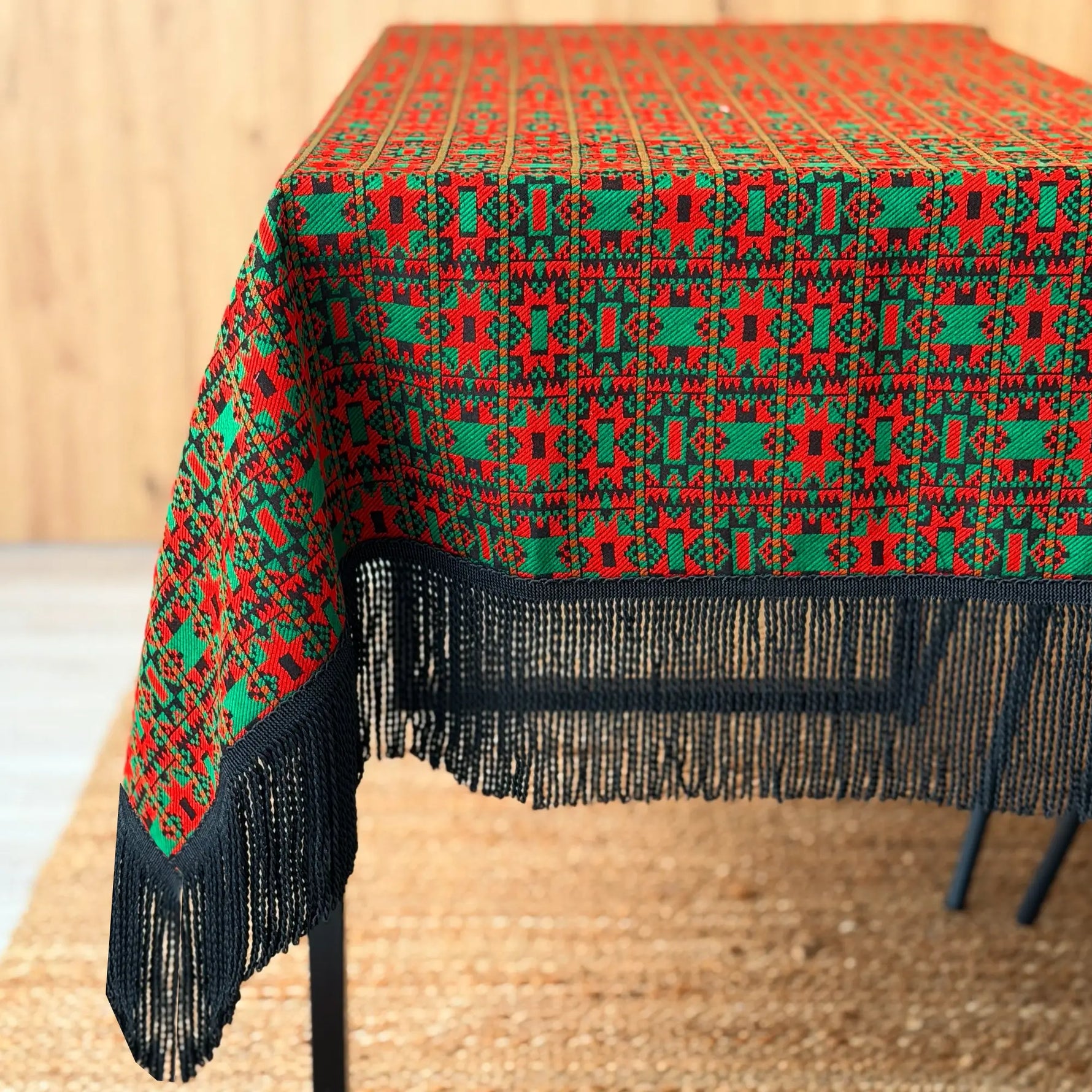 Tablecloth with red and green geometric pattern and black fringe on a wooden table.