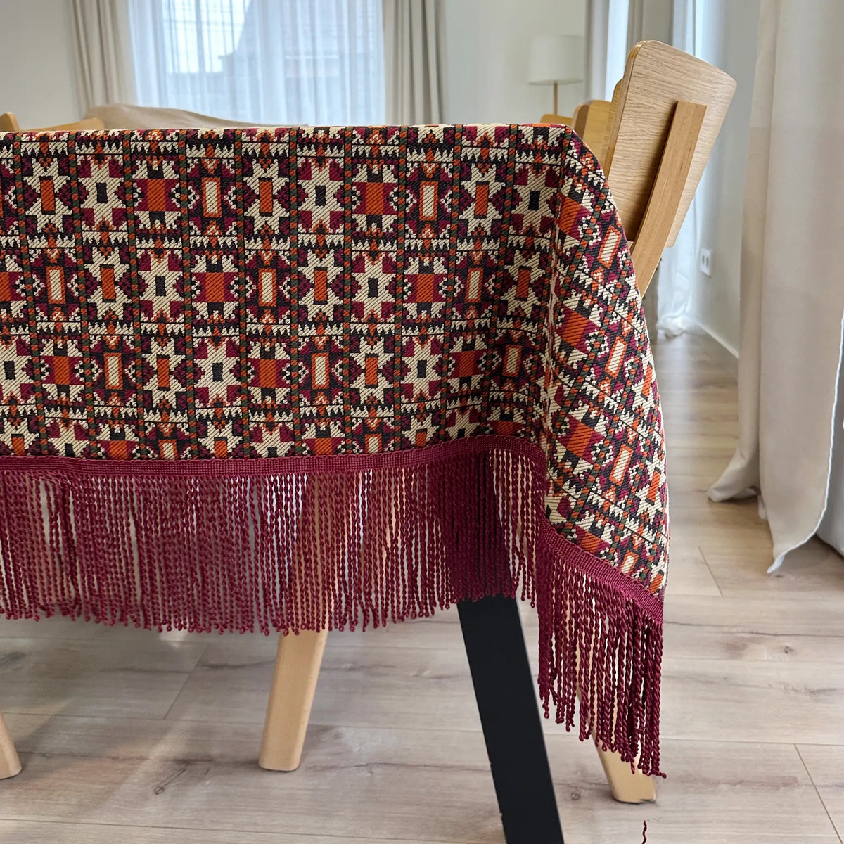 Patterned tablecloth with fringe on a wooden table in a room with curtains.