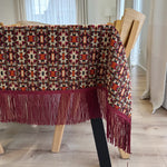 Patterned tablecloth with fringe on a wooden table in a room with curtains.