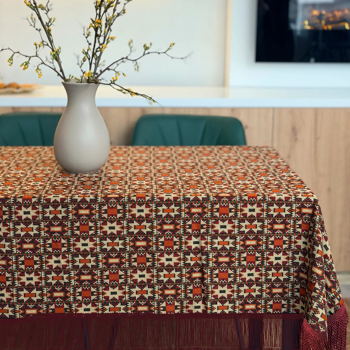 Table with a patterned tablecloth, vase with branches, and blurred background