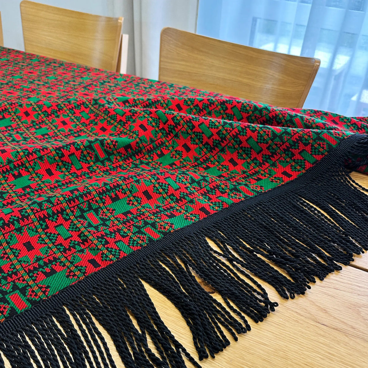 Red and green patterned throw blanket with black tassels on a wooden table.