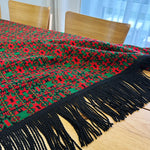 Red and green patterned throw blanket with black tassels on a wooden table.
