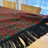Red and green patterned throw blanket with black tassels on a wooden table.