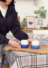 Person holding two gradient blue mugs on a woven mat with a home setting in the background.