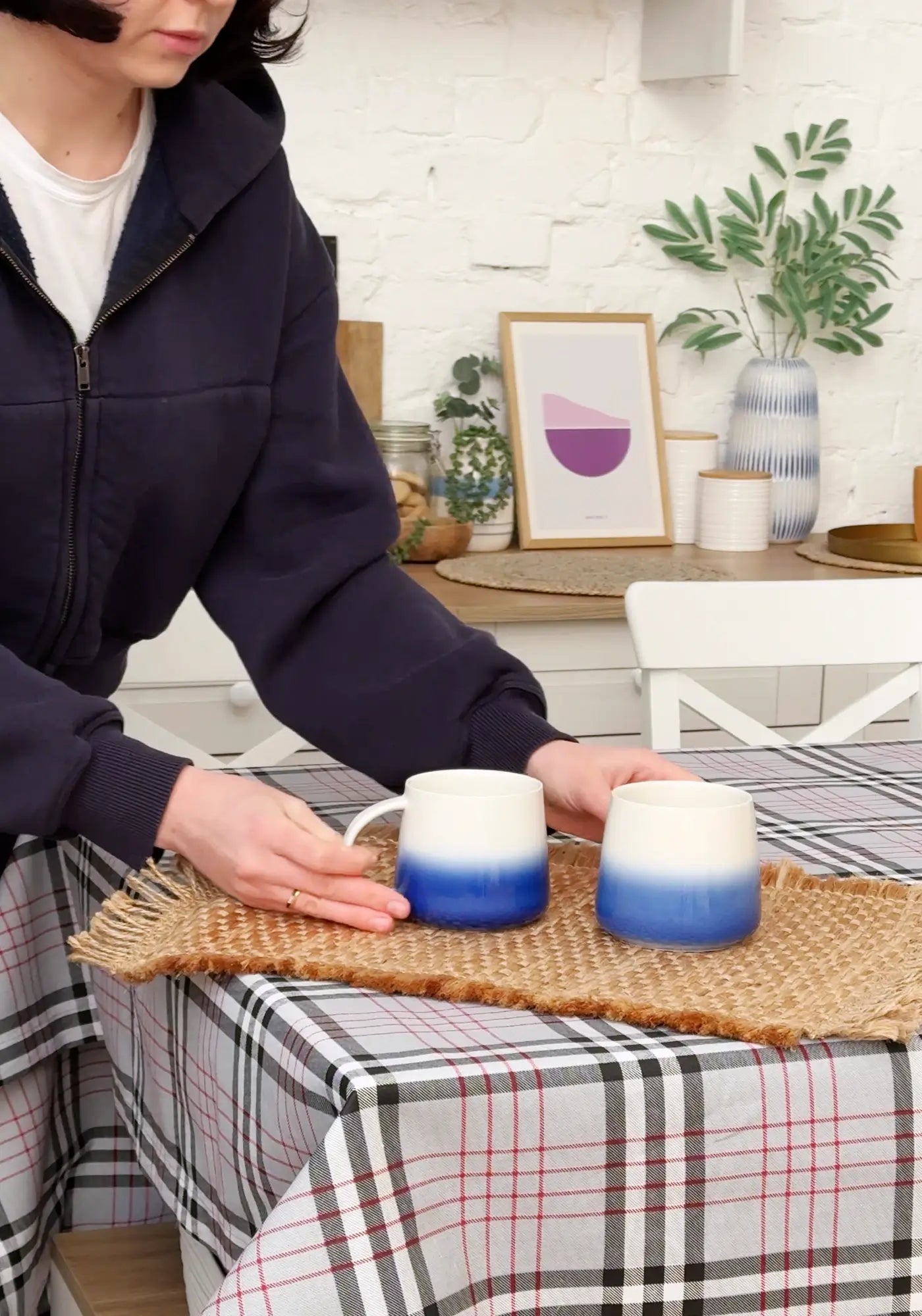 Person holding two gradient blue mugs on a woven mat with a home setting in the background.