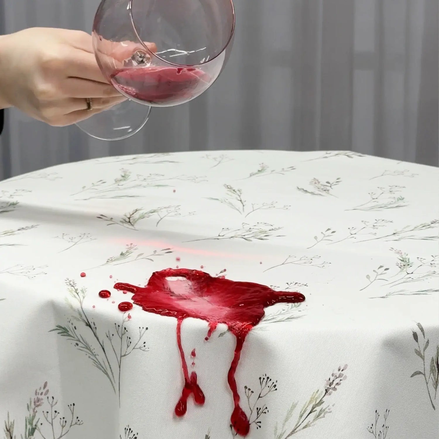Red wine spill on a floral-patterned tablecloth with a hand holding a glass above it.