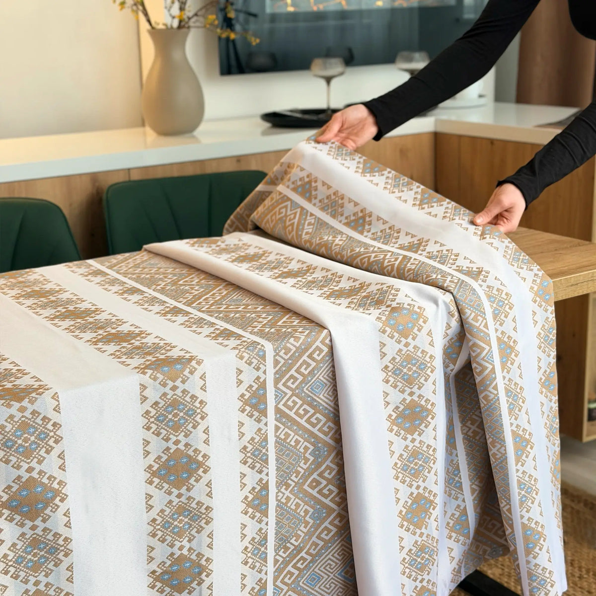 Person arranging patterned tablecloth on a dining table with a modern kitchen background.