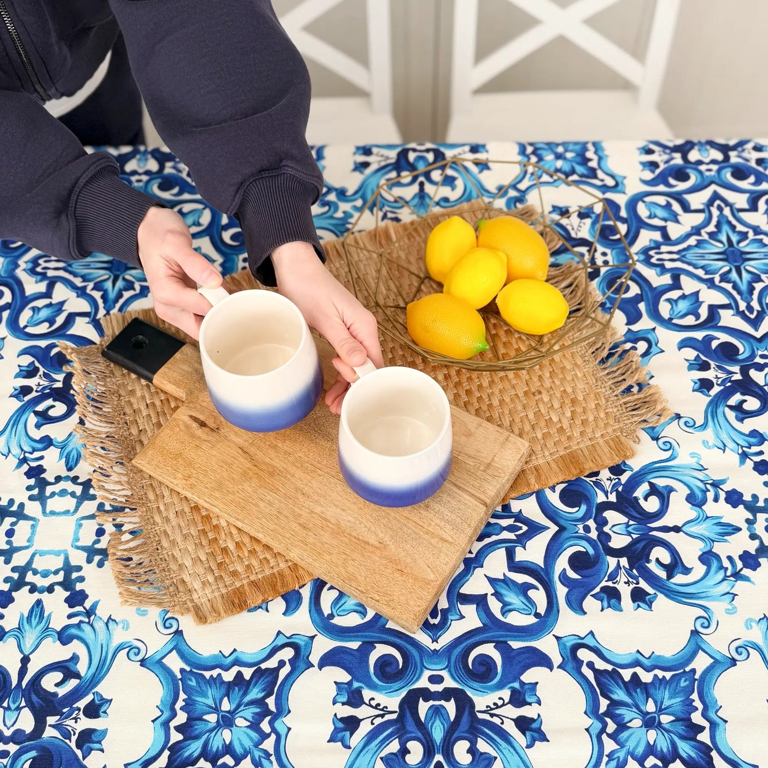 Two ceramic mugs with a gradient design on a woven tray with lemons, placed on a table with a blue and white patterned tablecloth.