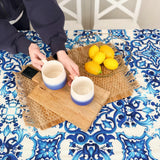 Two ceramic mugs with a gradient design on a woven tray with lemons, placed on a table with a blue and white patterned tablecloth.